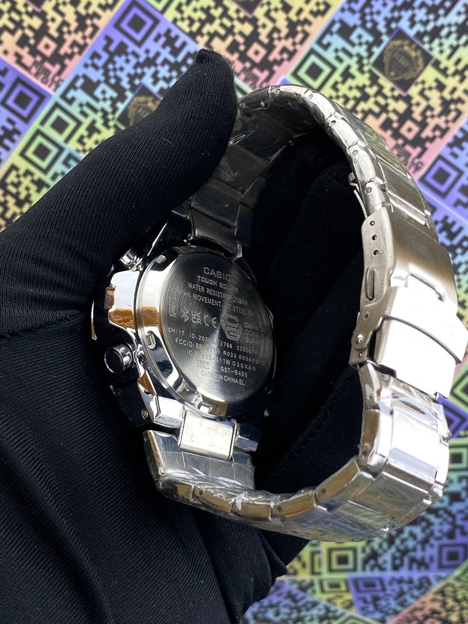 Silver Casio watch held by a gloved hand against a colorful geometric pattern background