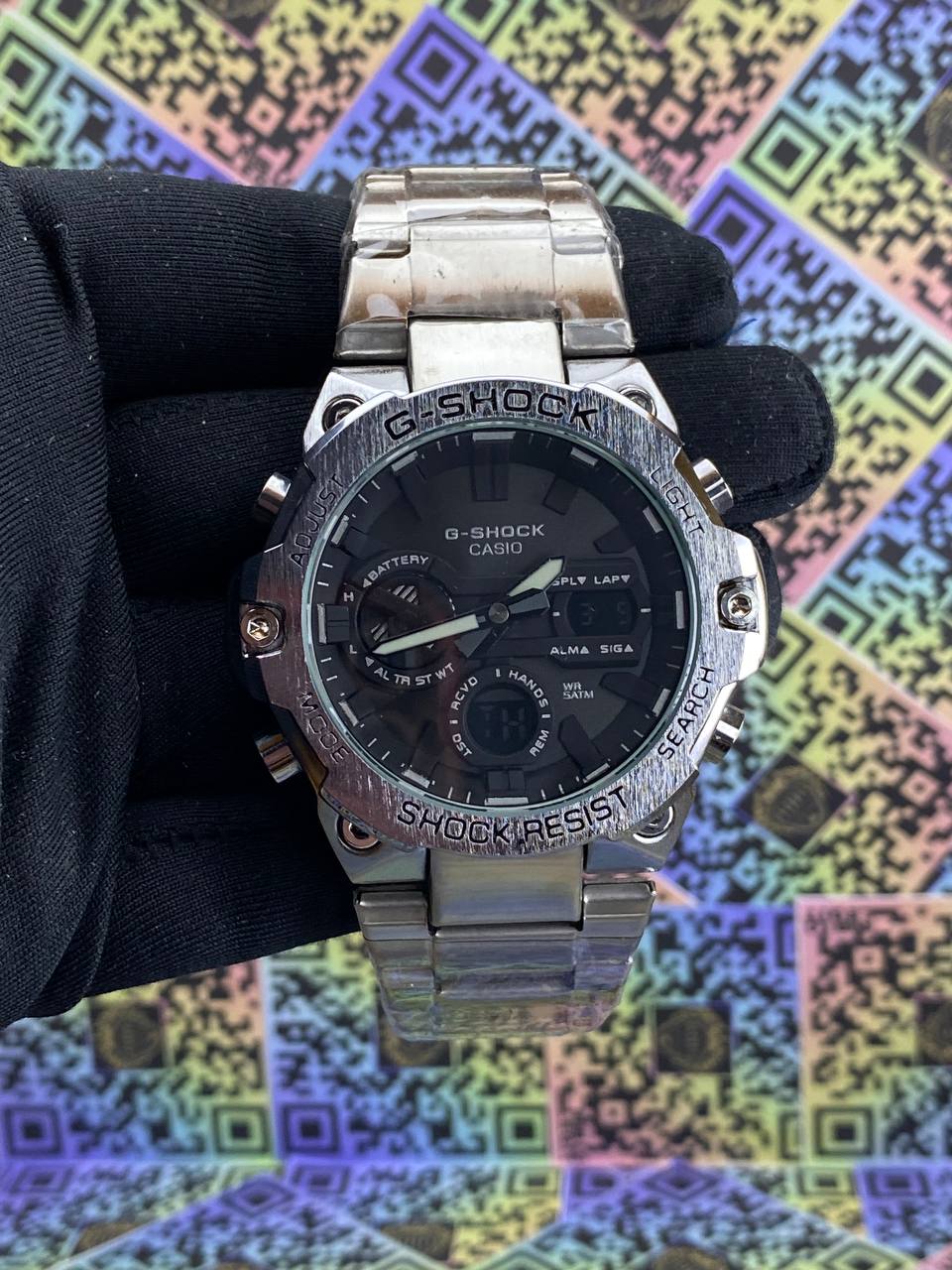 Silver Casio G-Shock watch held by a gloved hand against a colorful geometric pattern background.