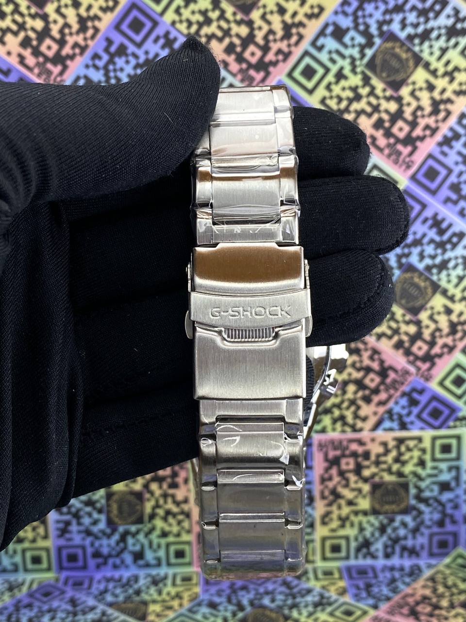 Silver G-Shock watch band held by a gloved hand against a colorful geometric patterned background