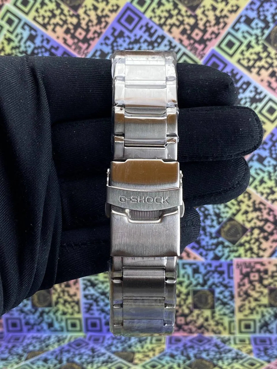 Silver G-Shock watch band held by a gloved hand against a colorful geometric patterned background