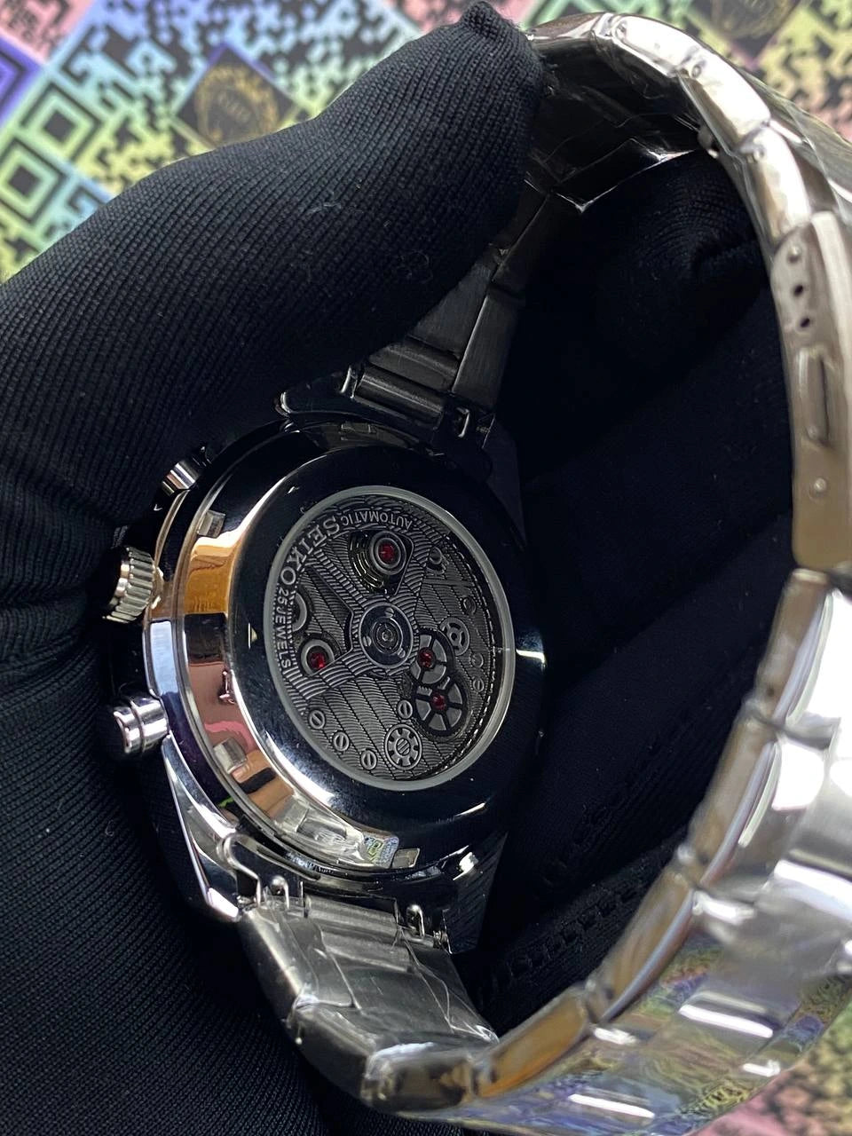 Close-up of a silver watch with visible mechanical components, held by a black gloved hand against a colorful geometric pattern.