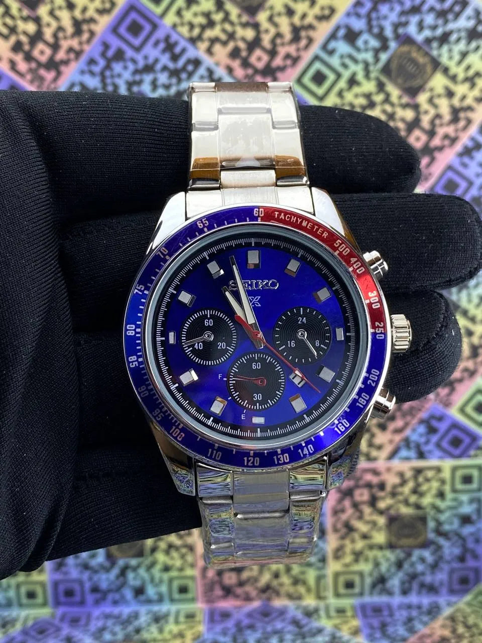 Silver watch with blue face held by a gloved hand against a colorful geometric pattern background